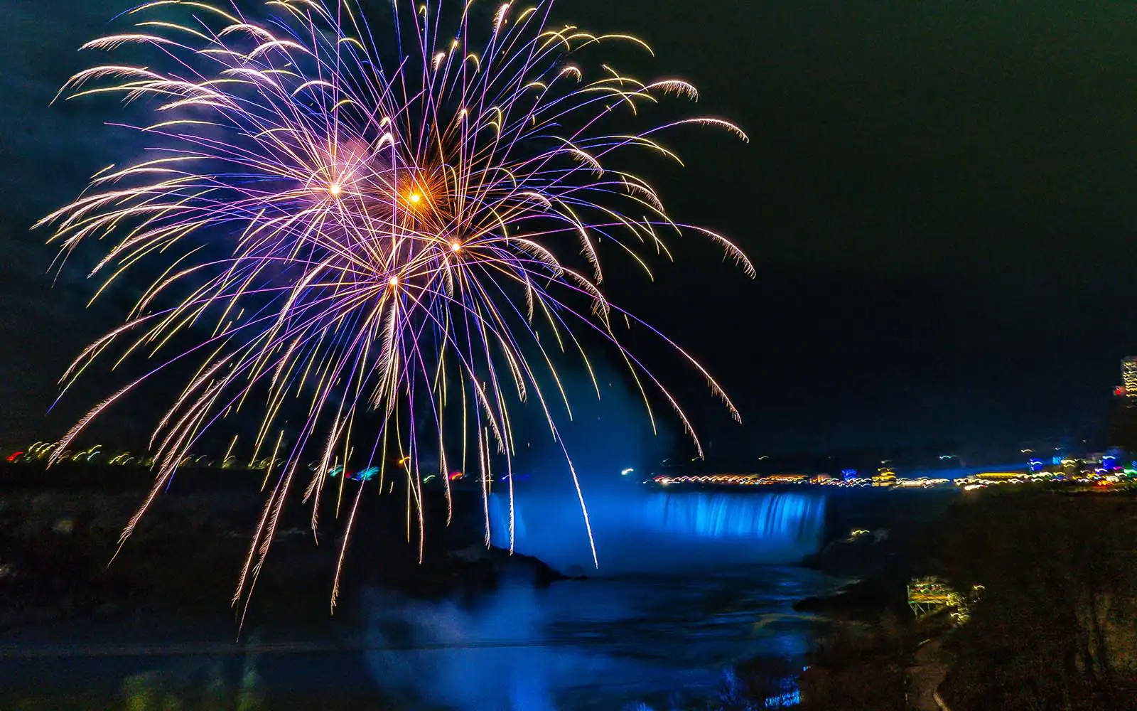 White, blue and purple fireworks bursting infront of the Canadian Horseshoe Falls illuminated in blue 