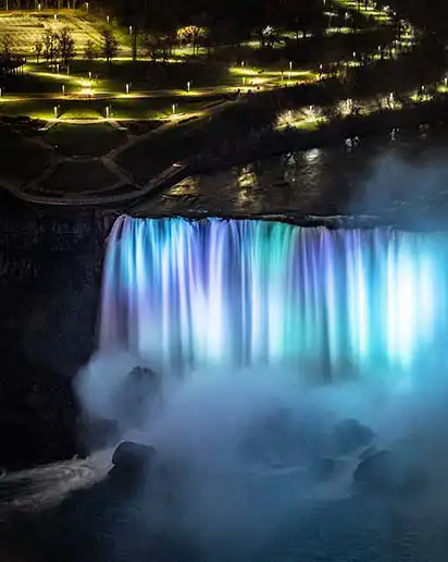 Nighttime view of Niagara Falls, brightly lit with water flowing under dark sky.