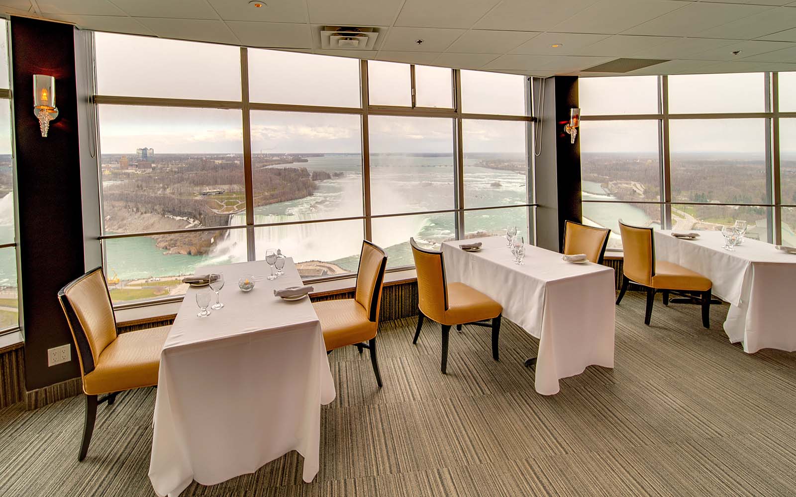 Restaurant with large windows overlooking Niagara Falls; tables are set with white tablecloths, brown chairs, and place settings.