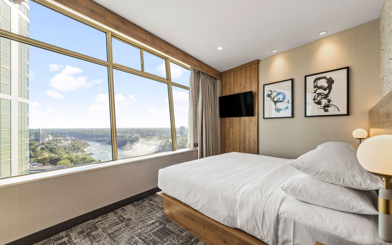 Hotel bedroom with a king-sized bed, light wooden headboard, abstract black-and-white wallpaper, a desk with a white countertop, gray-beige carpet, and soft, even lighting.