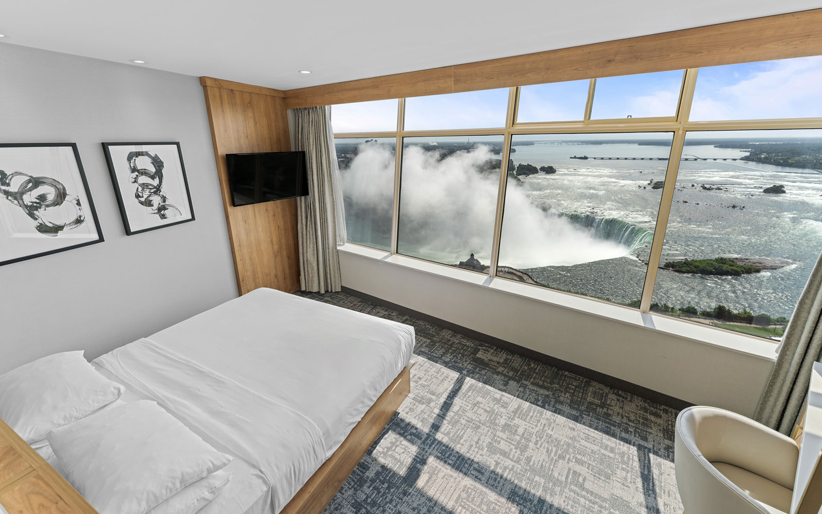 Modern hotel bedroom with a beige-gray carpet, wooden platform bed with white bedding, abstract art, large window with tree views, flat-screen TV, white vanity area, and golden shelving, all in a minimalist design.