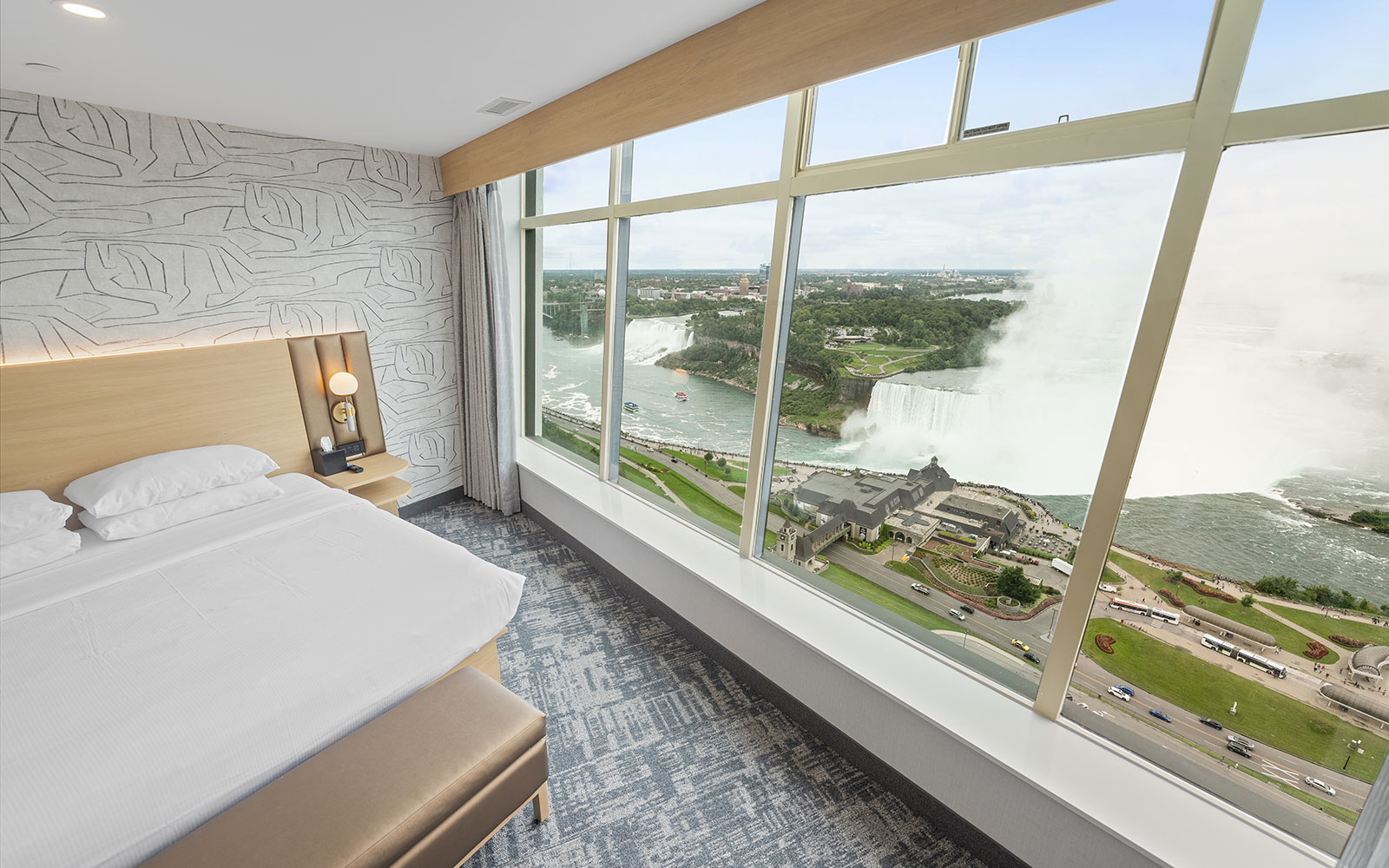 Hotel room with a large window showcasing Niagara Falls, featuring a neatly made bed and a built-in desk and wardrobe.