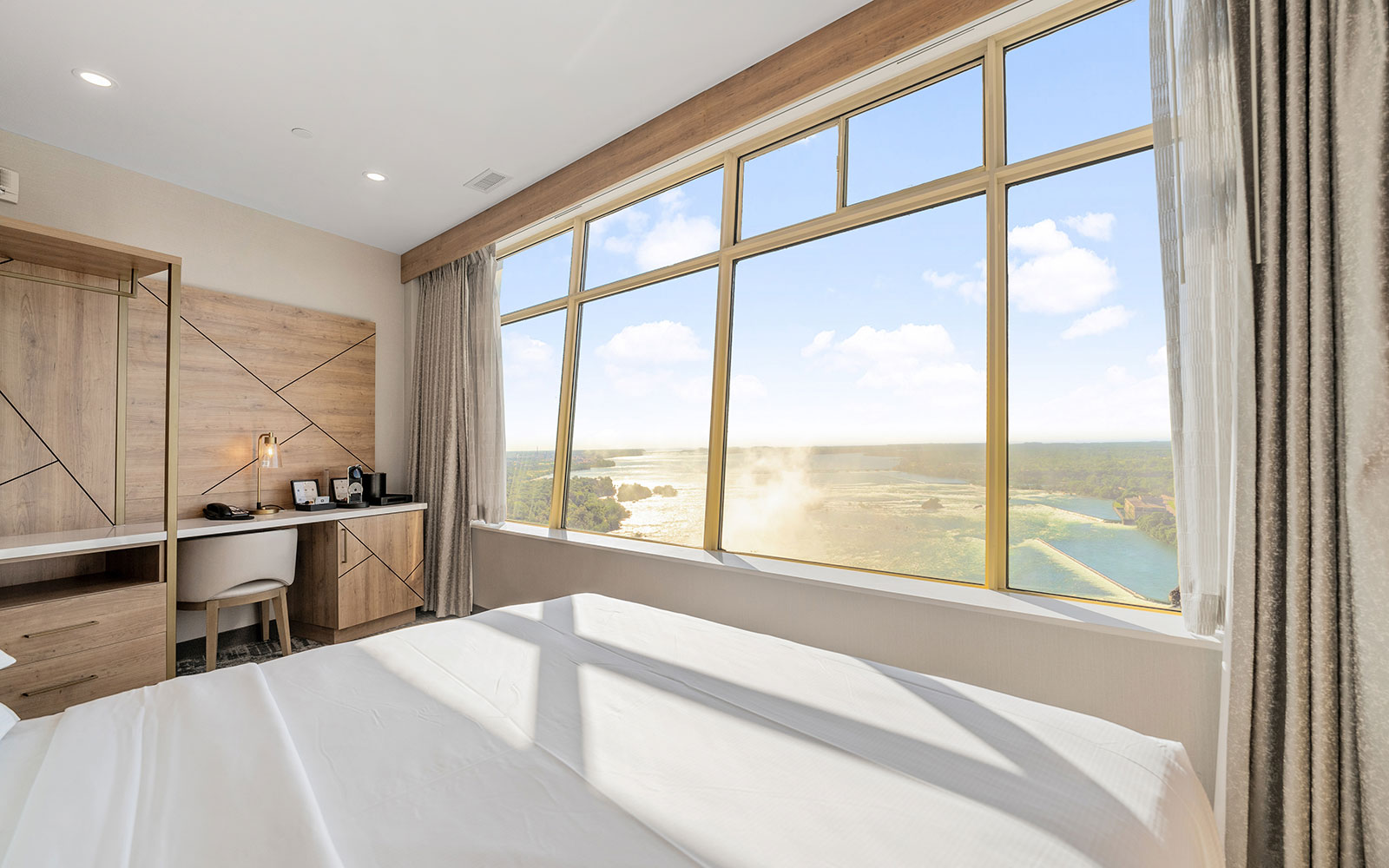 Hotel room with a large window offering a cityview, beige bed with white linens, marble-top desk, wooden wall unit, open wardrobe, and modern, airy design.